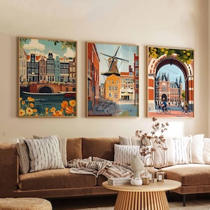 Pu&ograve; includere: Tre stampe artistiche incorniciate raffiguranti paesaggi urbani europei. L'opera presenta edifici colorati, canali, mulini a vento e ciclisti. Le stampe sono esposte sopra un divano marrone con cuscini a righe e un tavolino di legno.