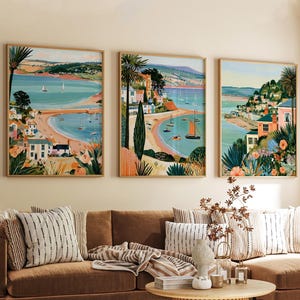Pode incluir: Um conjunto de três impressões de arte de parede emolduradas retratando paisagens costeiras com águas azul-turquesa, praias de areia, veleiros e casas coloridas entre palmeiras e flores vibrantes, num estilo ilustrativo detalhado.