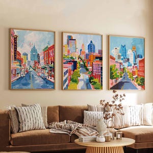 May include: Three framed cityscape paintings with a vibrant, colorful style. Each artwork depicts a street scene with buildings, roads, and a bright blue sky. The paintings are displayed above a brown sofa with decorative pillows.