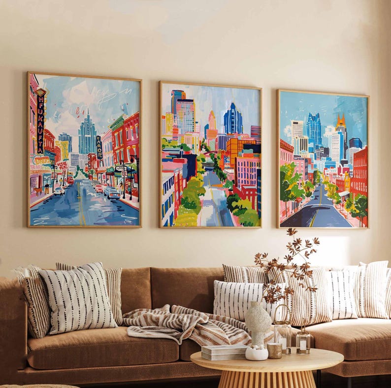 May include: Three framed cityscape paintings with a vibrant, colorful style. Each artwork depicts a street scene with buildings, roads, and a bright blue sky. The paintings are displayed above a brown sofa with decorative pillows.