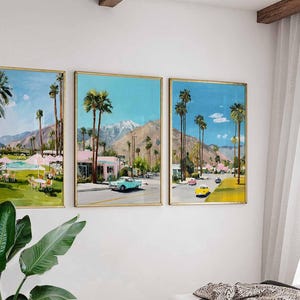May include: Set of three framed landscape paintings. Each painting depicts a sunny scene with palm trees, mountains, and vintage cars. The paintings have gold frames and are displayed on a white wall.