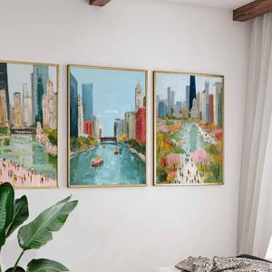 May include: Set of three framed cityscape paintings. Each artwork depicts a river scene with buildings, bridges, and people. The paintings feature a palette of blues, greens, pinks, and yellows, with gold frames. The artwork is displayed on a white wall.