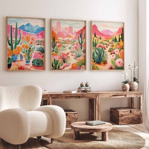 May include: Three framed prints of desert landscapes with cacti, flowers, and mountains in pink, green, and blue hues. The prints are hanging on a white wall above a wooden table and a white armchair.