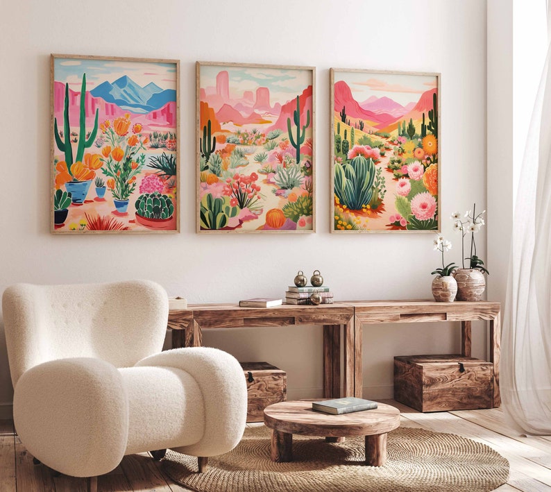 May include: Three framed prints of desert landscapes with cacti, flowers, and mountains in pink, green, and blue hues. The prints are hanging on a white wall above a wooden table and a white armchair.