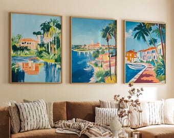 May include: Three framed landscape paintings with a coastal theme. Each features a vibrant, colorful depiction of buildings, palm trees, and water. The paintings are in wooden frames and hang above a brown sofa with striped pillows.