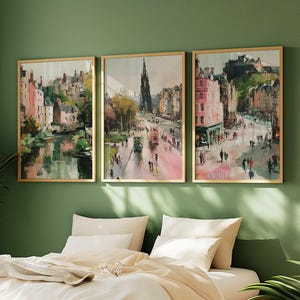 May include: Three framed landscape paintings with gold frames hang on a green wall above a bed. The paintings depict city scenes with buildings, streets, and people. The artwork has a watercolor style with pink, green, and brown tones.