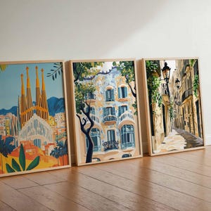 May include: Three framed art prints leaning against a wall. The prints depict architectural scenes, including a cathedral with tall spires, a building with ornate balconies, and a narrow street with streetlights. The frames are light-colored wood.