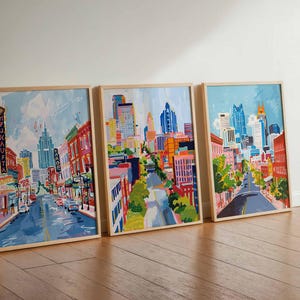 May include: Three framed city street art prints. Each features a colorful, impressionistic painting of a city street with buildings, cars, and trees. The artwork is in a wooden frame, leaning against a white wall.