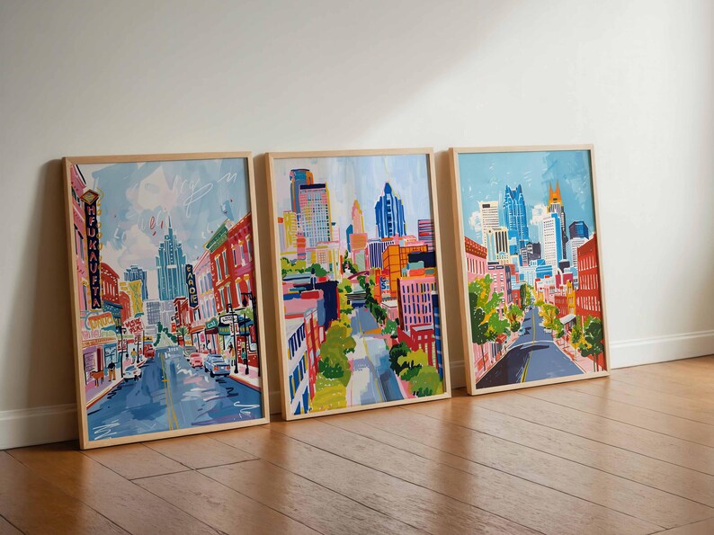 May include: Three framed city street art prints. Each features a colorful, impressionistic painting of a city street with buildings, cars, and trees. The artwork is in a wooden frame, leaning against a white wall.