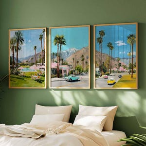 May include: Three framed art prints depicting a retro Palm Springs scene with palm trees, vintage cars, and pink buildings. The artwork is set against a blue sky and mountain backdrop, framed in gold, and displayed on a green wall.