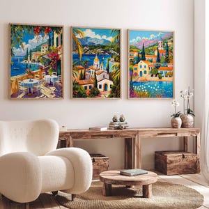 May include: Three colorful paintings of Mediterranean towns with blue water, white buildings, and lush greenery. The paintings are framed and hung on a white wall above a wooden table and a white armchair.