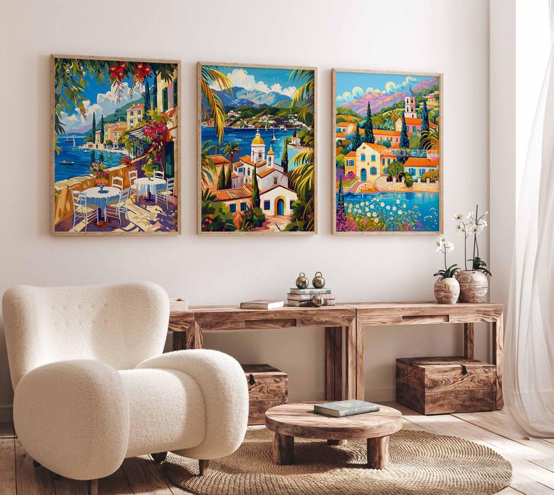 May include: Three colorful paintings of Mediterranean towns with blue water, white buildings, and lush greenery. The paintings are framed and hung on a white wall above a wooden table and a white armchair.