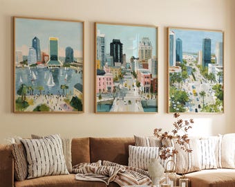 May include: Three framed cityscape paintings. The artwork depicts colorful buildings, roads, and water with sailboats. The paintings are in wooden frames and hang above a brown sofa with striped pillows.