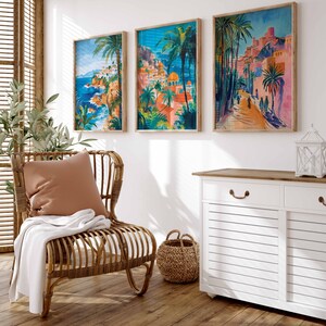 May include: Three framed prints of colorful, stylized paintings of Mediterranean townscapes with palm trees and blue water. The prints are hanging on a white wall above a white cabinet with a wooden top. A wicker chair with a brown cushion and a white throw blanket is in the foreground.