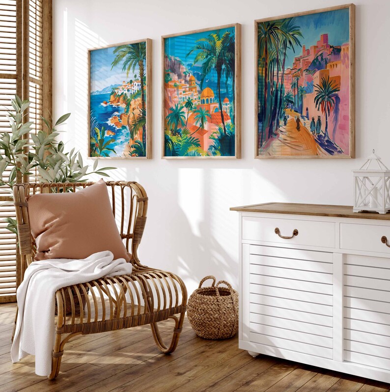 May include: Three framed prints of colorful, stylized paintings of Mediterranean townscapes with palm trees and blue water. The prints are hanging on a white wall above a white cabinet with a wooden top. A wicker chair with a brown cushion and a white throw blanket is in the foreground.