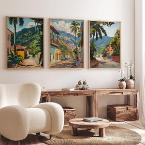 May include: Three framed landscape paintings depicting tropical scenes with palm trees, buildings, and mountains. The paintings are hung on a white wall above a wooden table and a white armchair.