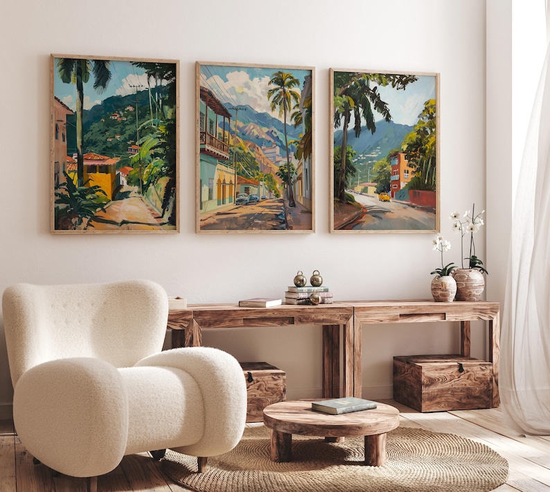 May include: Three framed landscape paintings depicting tropical scenes with palm trees, buildings, and mountains. The paintings are hung on a white wall above a wooden table and a white armchair.