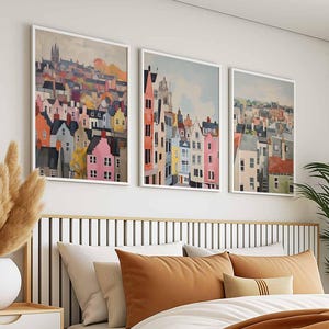 May include: Three framed art prints depicting colorful townscapes. The artwork features a variety of buildings in shades of pink, orange, yellow, and blue, set against a light sky. The prints are displayed above a bed with brown and white bedding.