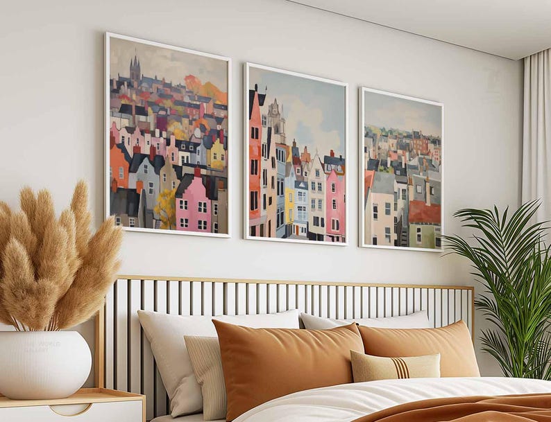 May include: Three framed art prints depicting colorful townscapes. The artwork features a variety of buildings in shades of pink, orange, yellow, and blue, set against a light sky. The prints are displayed above a bed with brown and white bedding.