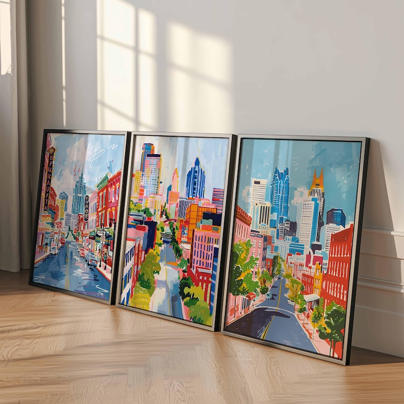 May include: Set of three framed cityscape art prints. Each print features a colorful, abstract depiction of a city street with buildings, trees, and a blue sky. The frames are black with silver inner edges, leaning against a white wall.