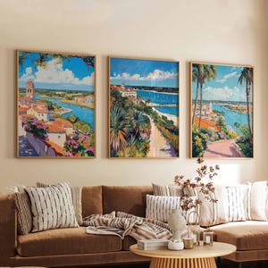 May include: Three framed landscape paintings showcasing coastal scenes with buildings, palm trees, and waterways. The artwork uses a vibrant colour palette of blues, greens, and oranges, with a focus on architectural details and natural elements. The paintings are displayed above a brown sofa.