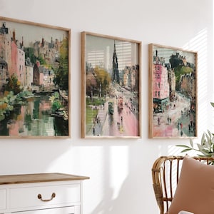 May include: Set of three framed art prints featuring impressionistic cityscapes. The artwork showcases buildings in shades of pink, green, and brown, with a focus on architectural details and street scenes. The frames are made of light-colored wood.