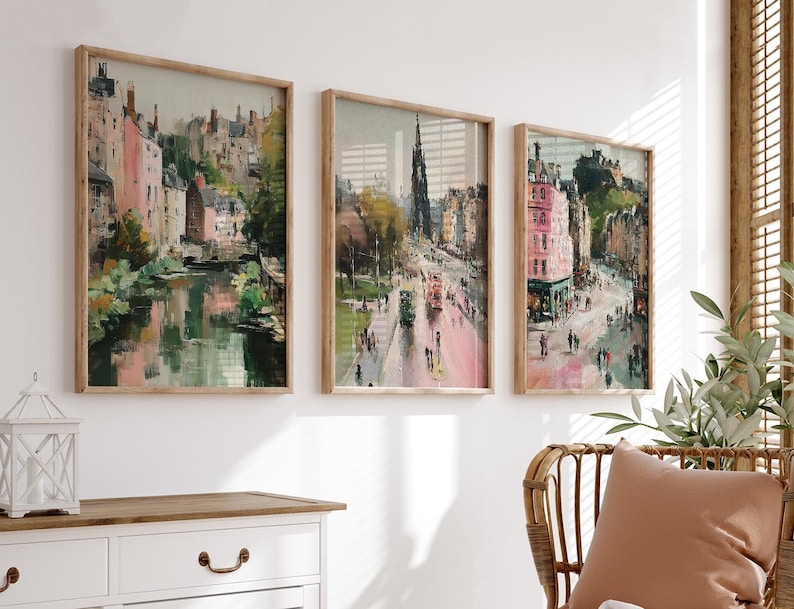 May include: Set of three framed art prints featuring impressionistic cityscapes. The artwork showcases buildings in shades of pink, green, and brown, with a focus on architectural details and street scenes. The frames are made of light-colored wood.
