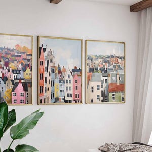 May include: Three framed art prints depicting colorful cityscapes. The artwork features buildings in shades of pink, orange, blue, and gray, set against a light blue sky. The frames are gold-colored, and the overall style is reminiscent of watercolor paintings.