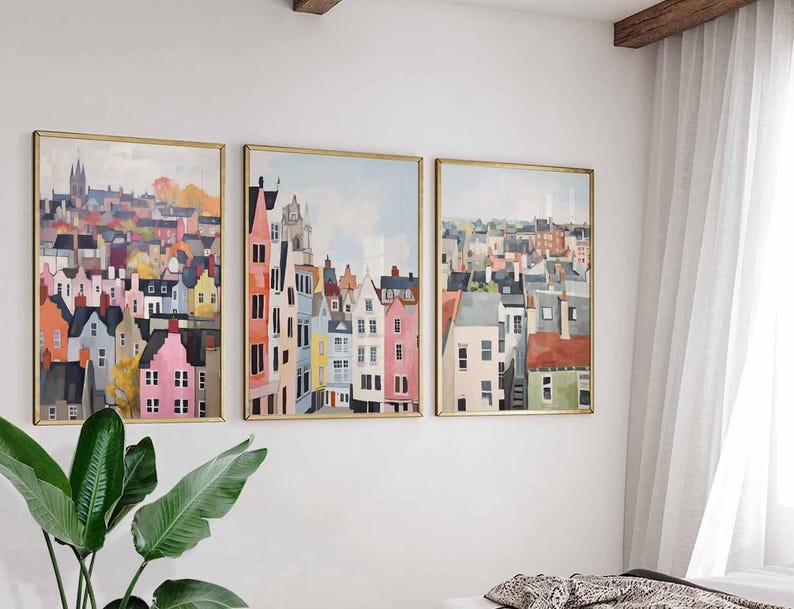 May include: Three framed art prints depicting colorful cityscapes. The artwork features buildings in shades of pink, orange, blue, and gray, set against a light blue sky. The frames are gold-colored, and the overall style is reminiscent of watercolor paintings.