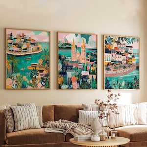 May include: Three framed art prints depicting colorful cityscapes with buildings, water, and lush greenery. The artwork features a vibrant palette of blues, greens, pinks, and yellows, creating a lively and artistic aesthetic. The prints are displayed above a brown sofa.