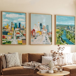 May include: Three framed cityscape paintings with wooden frames. The paintings depict colourful cityscapes with buildings, roads, and blue skies. The artwork is displayed above a brown sofa with striped cushions.