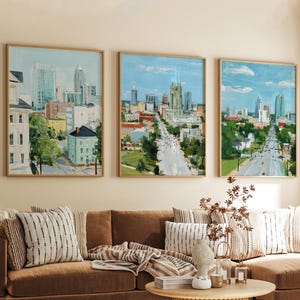 May include: Three framed cityscape paintings with gold-colored frames. The paintings depict a city street with buildings and trees. The color palette includes blues, greens, and browns. The paintings are displayed above a brown sofa with striped pillows.