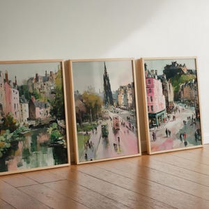 May include: Three framed watercolor paintings of a European city scene. The artwork depicts buildings, streets, and people, with a color palette of pink, green, and brown. The frames are made of light-colored wood.