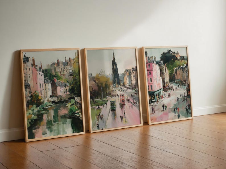 May include: Three framed watercolor paintings of a European city scene. The artwork depicts buildings, streets, and people, with a color palette of pink, green, and brown. The frames are made of light-colored wood.