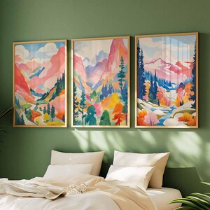 May include: Three framed landscape art prints. Each print features a colorful watercolor painting of a mountain scene with trees and a river. The artwork is in shades of pink, blue, green, yellow, and orange. The frames are gold.