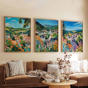 Op de afbeelding: Drie ingelijste landschapsschilderijen tonen een Europees dorp in een vallei, met kleurrijke bloemen op de voorgrond. Het kunstwerk heeft een levendig kleurenpalet van blauw, groen, oranje en roze, met een houten lijst.