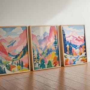 May include: Three framed landscape art prints. Each features a watercolor painting of a mountain scene with pink, blue, and green hues. The artwork is displayed on a wooden floor against a white wall.
