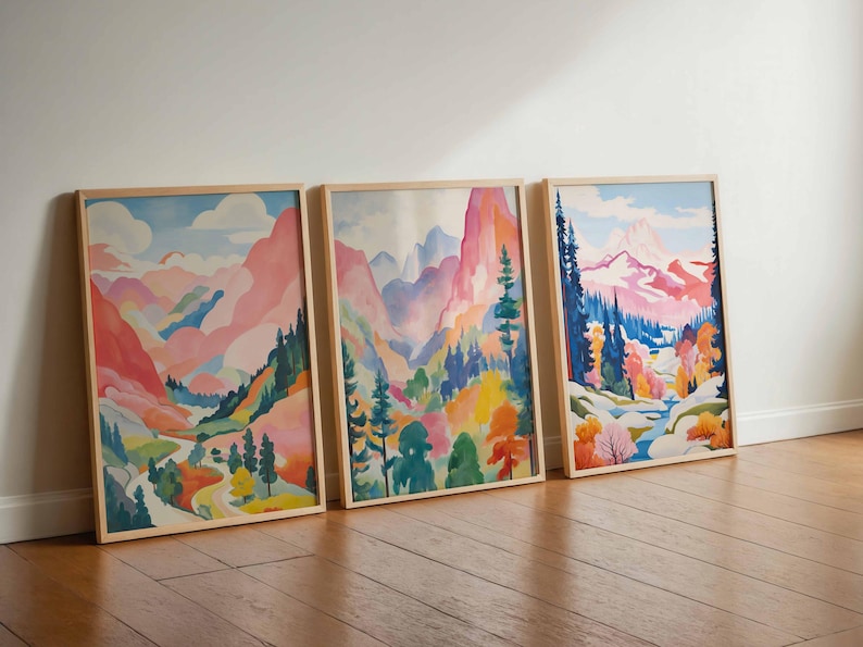 May include: Three framed landscape art prints. Each features a watercolor painting of a mountain scene with pink, blue, and green hues. The artwork is displayed on a wooden floor against a white wall.