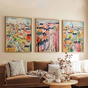 May include: Three framed watercolour paintings showcase vibrant townscapes with buildings, trees, and roads. The artwork uses pinks, oranges, blues, and greens. The paintings are displayed above a brown sofa with decorative cushions.