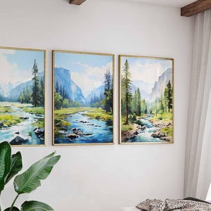 May include: Three framed landscape paintings depicting a river flowing through a valley with mountains and trees. The artwork features a watercolor style with shades of blue, green, and gold. The paintings are displayed on a white wall.