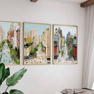 May include: Three framed cityscape paintings. Each features a street scene with tall buildings, trees, and yellow taxis. The artwork has a gold frame and is mounted on a white wall. The paintings are in a row, creating a cohesive display.