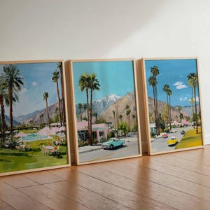 May include: Three framed landscape paintings depicting a sunny street scene with palm trees, mountains, and vintage cars. The artwork features a vibrant color palette with blue skies, green grass, and pink buildings. The frames are made of light-colored wood.