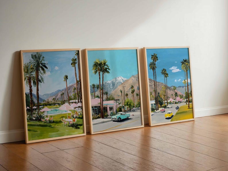 May include: Three framed landscape paintings depicting a sunny street scene with palm trees, mountains, and vintage cars. The artwork features a vibrant color palette with blue skies, green grass, and pink buildings. The frames are made of light-colored wood.