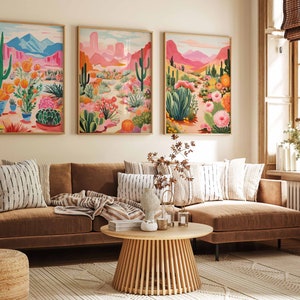 May include: Three framed illustrations of desert landscapes with cacti, flowers, and mountains in shades of pink, green, and blue. The illustrations are hanging on a wall above a brown sectional sofa with striped pillows.