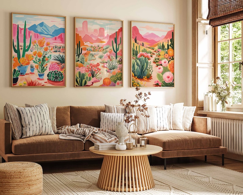 May include: Three framed illustrations of desert landscapes with cacti, flowers, and mountains in shades of pink, green, and blue. The illustrations are hanging on a wall above a brown sectional sofa with striped pillows.