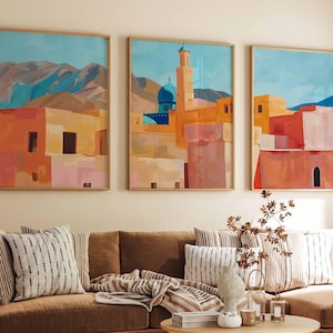 May include: Three framed abstract art prints depicting a cityscape with buildings in shades of orange, pink, and yellow, set against a blue sky and mountains. The artwork is displayed above a brown sofa with striped pillows.