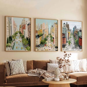 May include: Three framed cityscape paintings. Each features a street scene with tall buildings, trees, and yellow taxis. The artwork has a light color palette with a focus on the buildings and the road. The frames are a light wood color.