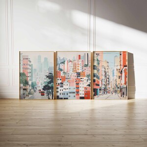 May include: Three framed art prints depicting a cityscape with colorful buildings, a street, and people. The prints are in a light blue, pink, and yellow color scheme. The prints are arranged in a row on a light brown wood floor.