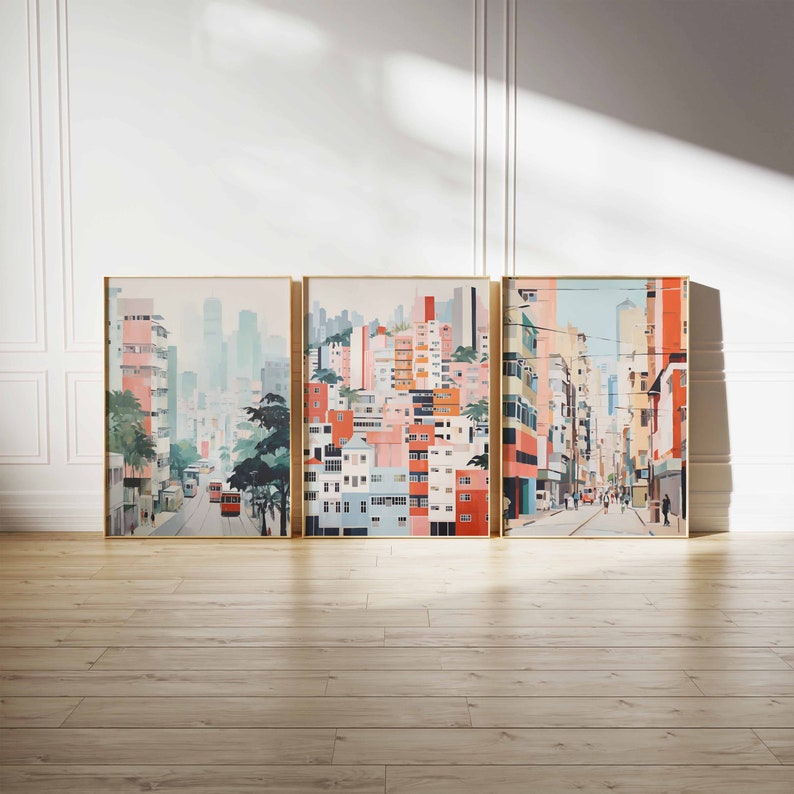 May include: Three framed art prints depicting a cityscape with colorful buildings, a street, and people. The prints are in a light blue, pink, and yellow color scheme. The prints are arranged in a row on a light brown wood floor.