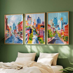May include: Three framed cityscape paintings with gold frames hang above a bed. The artwork depicts colorful city streets with buildings, trees, and cars. The paintings are in shades of blue, red, orange, and yellow.